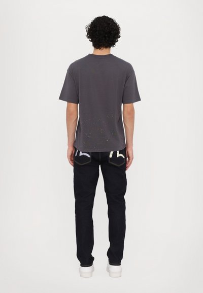 Una camiseta de algodón gris de mangas cortas con un ligero diseño salpicado, combinada con unos jeans azul oscuro y zapatillas blancas, vista desde la parte posterior.