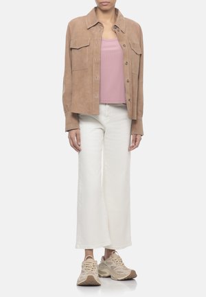 Persona che indossa una giacca beige in camoscio con bottoni, canotta rosa, pantaloni bianchi a gamba larga e scarpe da corsa beige, in piedi su sfondo bianco.