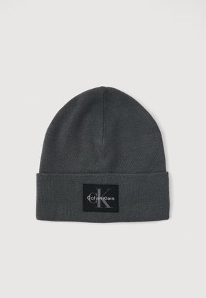 Szürke kötött sapka lehajtható peremmel, fekete négyzet alakú emblémával, rajta fehér "Calvin Klein" logóval és stilizált "CK" jellel.