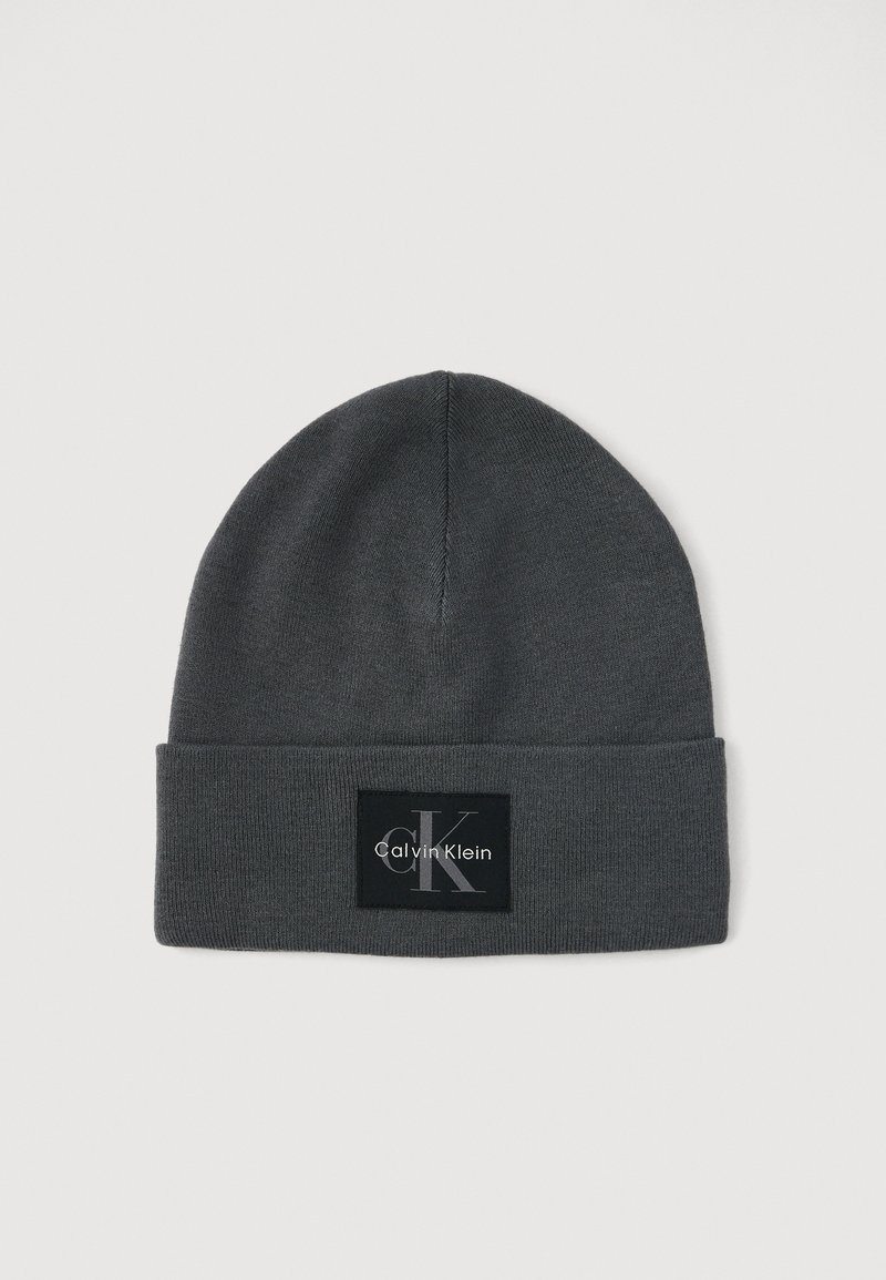 Harmaa neulehattu, jossa on käännetty reuna. Hatun etuosassa on musta neliöpatch, jossa valkoinen "Calvin Klein" -logo ja tyylitelty "CK" -embleemi.