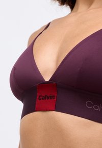 Sclipici cu decolteu în V, de culoare purpurie profundă, cu un material neted, având o bandă largă cu o etichetă roșie pe care este înscris "Calvin" cu litere negre.