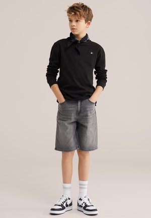 Niño con camisa negra de manga larga, pañuelo negro en la cabeza, pantalones cortos de mezclilla gris, calcetas blancas y zapatillas blancas y negras, de pie con las manos en los bolsillos.
