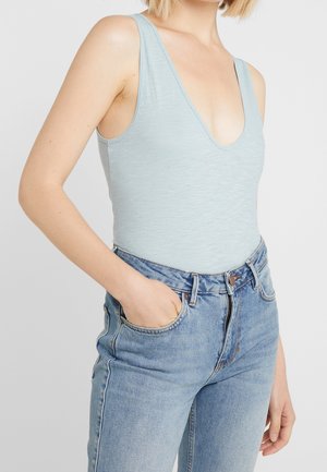 Femme portant un débardeur bleu clair à encolure dégagée, rentré dans un jean bleu taille haute, avec une main dans la poche du jean.
