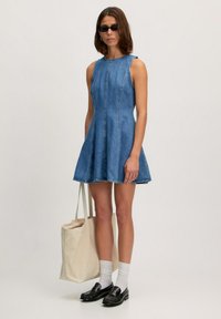Denim mouwloze jurk met een uitlopende silhouette, lichtblauwe kleur, zichtbare stiksels en een rafelige zoom. Model draagt een beige tote bag en heeft zwarte instappers met witte sokken aan.
