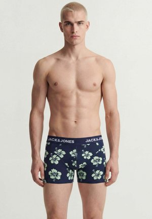 Jeune homme aux cheveux blonds courts portant un boxer bleu avec imprimé floral blanc et ceinture de marque, debout devant un fond uni.