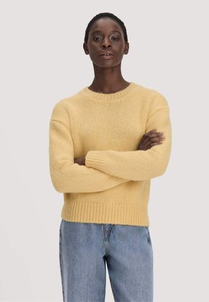 Persoon met kort haar die een geel gebreide trui en blauwe jeans draagt, met gekruiste armen staand tegen een effen lichte achtergrond.