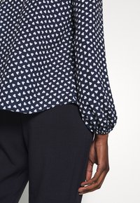 Blouse marine à motif cœurs blancs, avec des manches froncées. Associée à un pantalon sombre, affichant un tissu texturé.