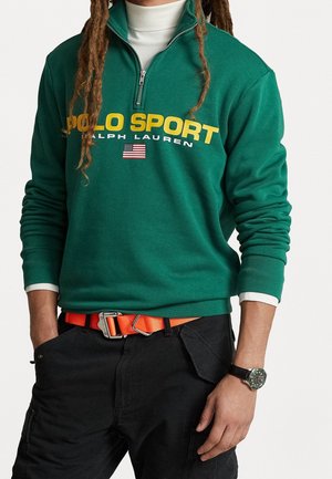 Person trägt einen grünen Polo Sport Ralph Lauren Pullover mit Viertelreißverschluss, einen weißen Rollkragenpullover, schwarze Hosen mit orangefarbenem Gürtel und eine schwarze Armbanduhr.