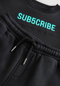 Sort sweatshirt og joggers med en glat tekstur. Sweatshirten har "SUB5CRIBE" i turkise bogstaver på ryggen og en elastisk talje.
