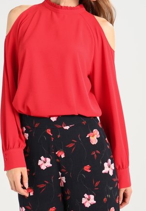 Kvinde iført en rød cold-shoulder top kombineret med sorte højhøjede bukser med et pink og rødt blomsterprint.