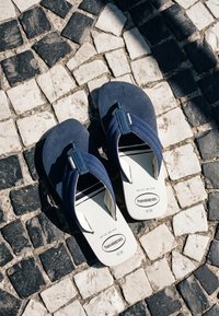 Ζευγάρι ναυτικό μπλε και λευκά σανδάλια Havaianas σε μαύρο-άσπρο, μοτίβο λιθόστρωτο κάτω από έντονο ηλιακό φως.