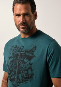 Camiseta de algodón color teal con un destacado diseño gráfico negro de dos figuras con barba. Las mangas cortas y el cuello redondo completan el diseño.