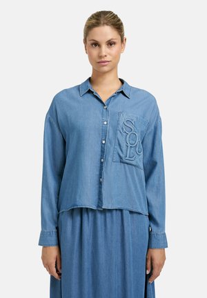 Frau trägt ein hellblaues Denim-Hemd mit Knöpfen und großer Brusttasche, auf der die Buchstaben "SOLO" geprägt sind, sowie einen passenden Rock.