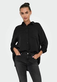 Camisa negra de botones confeccionada con un tejido texturizado, con un bolsillo en el lado izquierdo, combinada con jeans ajustados de color gris oscuro.