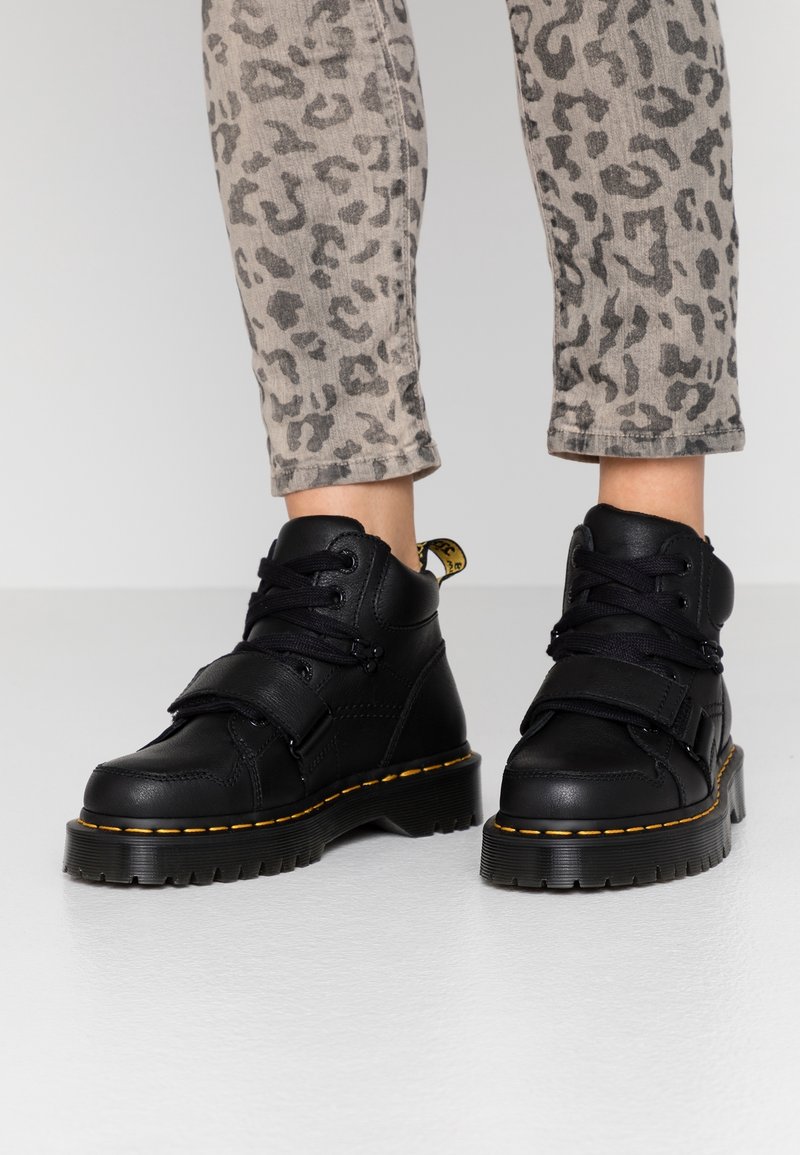 Černé kožené kotníkové boty se šněrováním a páskem, s masivní podrážkou a žlutým prošíváním. Nosené s šedými kalhotami s leopardím vzorem.