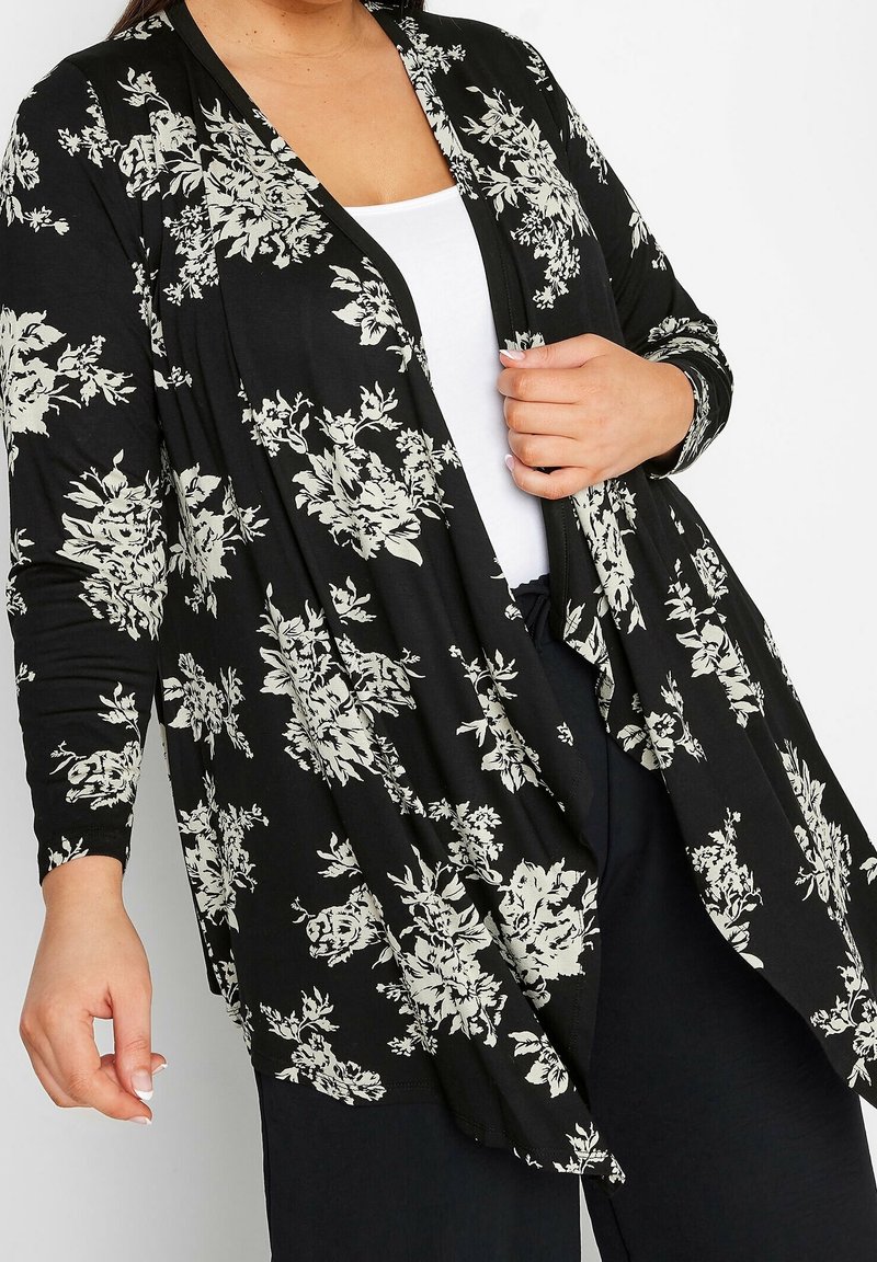 Femme portant un cardigan noir à manches longues ouvert sur le devant avec un motif floral blanc, par-dessus un haut blanc et un pantalon noir, tenant le revers du vêtement d'une main.