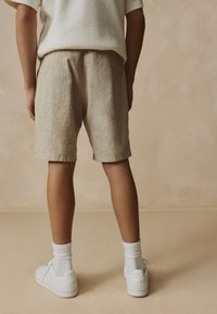 Beige linen shorts with a relaxed fit, featuring a waistband and rolled hems, paired with white sneakers and socks. Plain background.