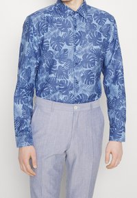 Ljusblå skjorta med knappar som har ett tropiskt bladmönster i mörkblått. Matchas med ljusgrå skräddarsydda byxor.