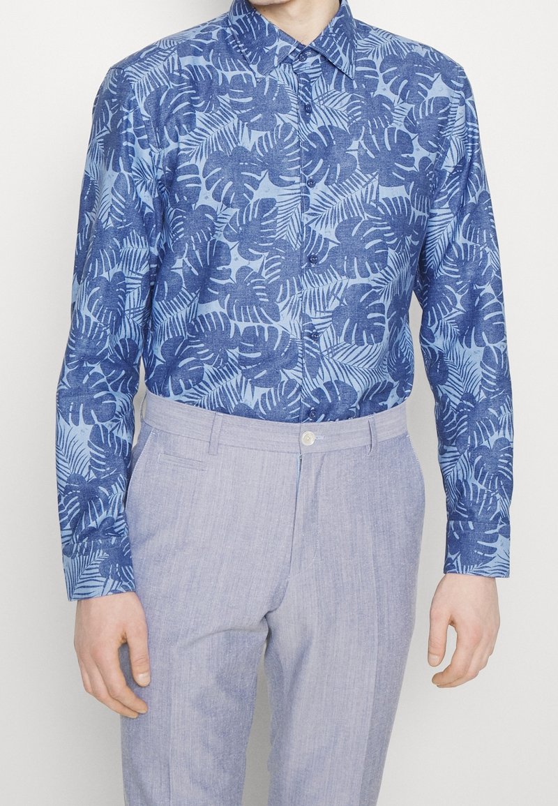 Ljusblå skjorta med knappar som har ett tropiskt bladmönster i mörkblått. Matchas med ljusgrå skräddarsydda byxor.