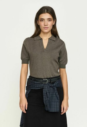 Donna con capelli lisci castani indossa una camicia marrone a maniche corte con colletto, una gonna nera e una camicia a quadri blu annodata in vita, frontale.