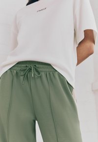 T-shirt bianco con stampa "PARIS", abbinato a pantaloni della tuta verde oliva con vita elasticizzata e tasche laterali. Tessuto morbido e liscio.