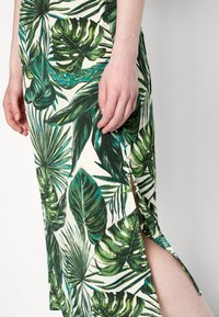 Robe à imprimé floral présentant de grandes feuilles vertes sur un fond crème. Elle possède une fente sur le côté, une coupe ajustée et un tissu doux et texturé.