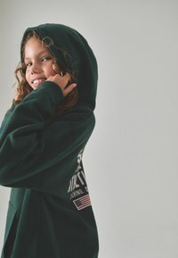 Felpa con cappuccio verde scuro con tasca frontale, caratterizzata da un testo bianco e una grafica della bandiera americana sul retro. Tessuto morbido, vestibilità comoda e design con cappuccio.