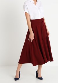 Jupe midi bordeaux avec une texture lisse, détail de taille nouée et une silhouette évasée, accompagnée d'une chemise blanche et de talons navy.