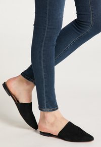 Schwarze Wildleder-Mules mit spitzem Zeh und offenem Rücken, kombiniert mit eng anliegenden blauen Jeans, die eine glatte Textur und ein klares Design zeigen.