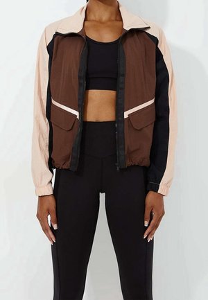Mujer con una chaqueta combinada en marrón, beige y negro sobre un sujetador deportivo negro y leggings negros de talle alto.