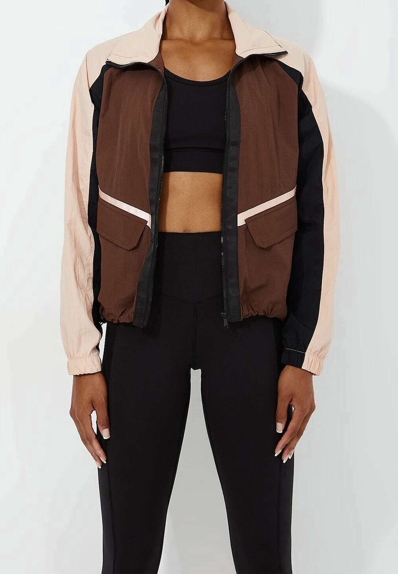 Femme portant une veste bicolore marron, beige et noire sur un soutien-gorge de sport noir et un legging noir taille haute.