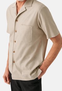 Camicetta beige a maniche corte con una tessitura strutturata, tre bottoni in legno e una tasca sul petto; colletto curato e vestibilità rilassata.