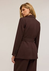 Blazer brown su misura realizzato in tessuto strutturato con vita cintata, maniche lunghe e polsini con tre bottoni, dotato di rever.