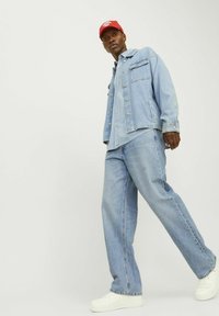 Lichtblauwe denimjacket met twee voorzakken en zilveren knopen, gecombineerd met bijpassende wijde spijkerbroek en witte sneakers. Rode pet op het hoofd.