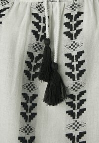 Prenda blanca con bordado geométrico negro que presenta borlas. Textura suave, cordón con dos borlas negras en el escote.
