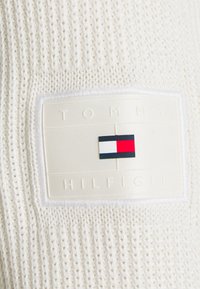 Tessuto lavorato a maglia bianco con una superficie texture. Presenta un'etichetta rettangolare Tommy Hilfiger con un design a blocchi di colore in rosso, bianco e blu.