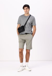 T-shirt en coton gris avec logo, shorts beiges et sac bandoulière noir. Le modèle porte des baskets blanches et des chaussettes ; look décontracté avec l'ourlet des shorts roulé.