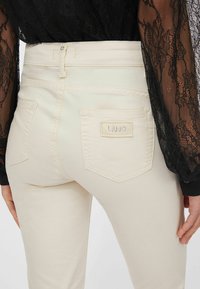 Vaalean kermaan vivahtavat farkut, joissa on istuva siluetti, kaksi takataskua ja pieni logokirjain. Kangas on sileä.