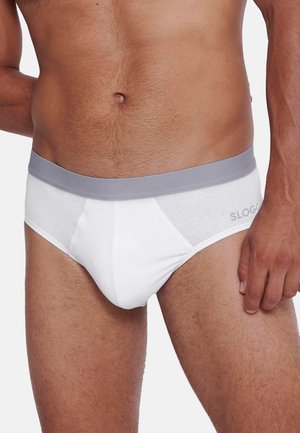 Torso maschile che indossa slip bianchi con fascia grigia in vita, in piedi contro uno sfondo chiaro e uniforme.