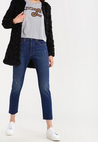 Zwart bontachtige jas over een grijs cropped t-shirt met oranje logo, gecombineerd met blauwe versleten jeans en witte sneakers.