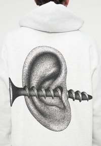 Γκρι φούτερ με κουκούλα που απεικονίζει ένα μαύρο γραφικό ενός αυτιού με βίδα που το διαπερνά, παρουσιάζοντας περίπλοκα μοτίβα από κουκκίδες και λεπτομέρειες.