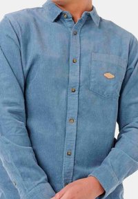 Camisa de pana azul con cuello abotonado, que presenta un bolsillo en el pecho y detalles en botones marrones. La tela tiene una textura acanalada vertical.