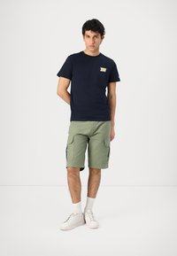 T-shirt navy con logo, pantaloni cargo verde oliva, sneakers bianchi e calzini corti bianchi. Texture del tessuto liscia; design casual.