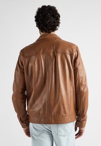 Man met krullend donker haar, gekleed in een bruin leren jack en lichtblauwe jeans, van achter gezien tegen een effen achtergrond.