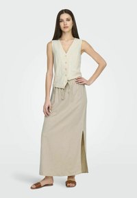 Chaleco de lino beige claro con escote en V y cierre de botones, combinado con una falda larga beige a juego con una abertura lateral. Sandalias marrones de tipo slip-on.