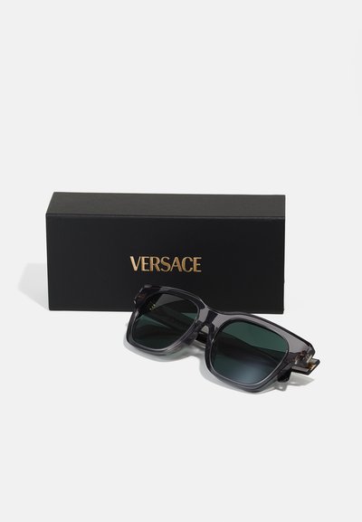 Vierkante zonnebril met donker groene lenzen en een doorschijnend grijs montuur. Wordt geleverd in een zwarte doos met gouden "Versace" logo.