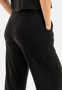 Pantalon large noir en tissu texturé avec un léger éclat. Comprend des poches latérales et des lignes fluides et ajustées.