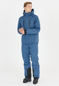 Mann trägt blaue Winterjacke und Hose mit schwarzen Skischuhen, steht und schaut seitlich vor weißem Hintergrund.