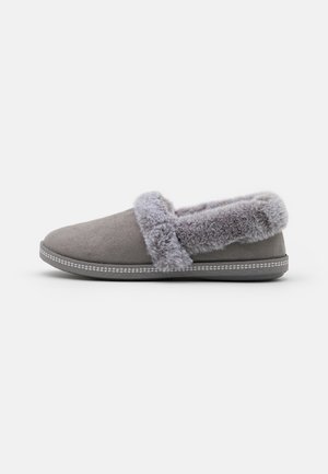 Grijze suède pantoffel met een zachte grijze bontvoering en -afwerking, met een platte zool en een decoratieve stiknaad rond de basis.