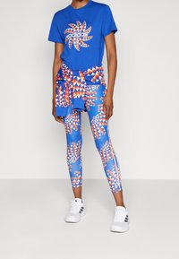 Modrá Adidas tričko s geometrickým vírovým logem, doplněné barevnými legínami s potiskem v modré, oranžové a bílé barvě. Bílé sportovní boty.